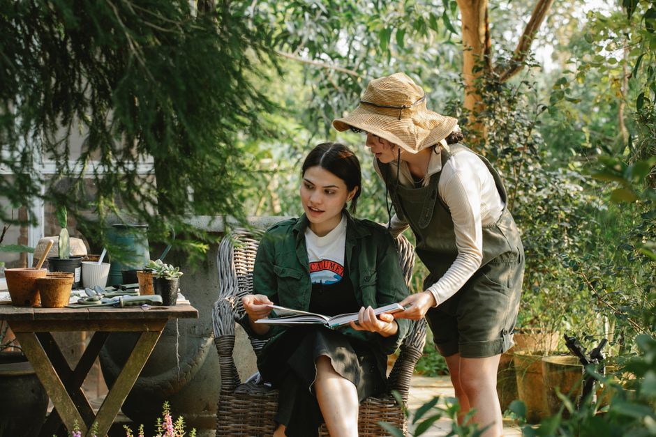 A woman wearing a large straw hat, beige long-sleeved top, and green gardening overalls bends over to show a booklet to a younger woman seated in a wicker chair in a lush garden setting. The seated wo