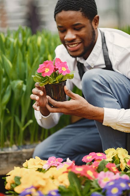A woman wearing a large straw hat, beige long-sleeved top, and green gardening overalls bends over to show a booklet to a younger woman seated in a wicker chair in a lush garden setting. The seated wo